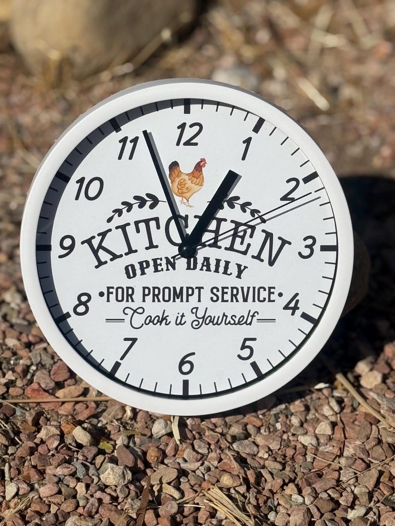 May include: White round clock with black hands and numbers. The clock face has a black and white illustration of a chicken and the words "Kitchen Open Daily" with the text "For Prompt Service - Cook it Yourself -" below.
