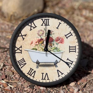 May include: A black framed clock with Roman numerals and a floral design featuring a white bathtub with black feet. The clock face has a vintage aesthetic.