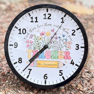 May include: A black and white clock with a colorful floral design and the word "TEACHER" in rainbow letters. The clock also has the text "Teach them, love them, watch them grow" and "Mrs. Rosenblum".