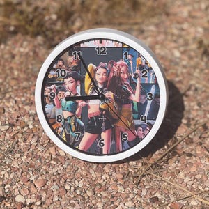 May include: A round wall clock with a white frame. The clock face features a collage of images of three women in various outfits. The clock hands are black. The clock is sitting on a bed of small brown rocks.