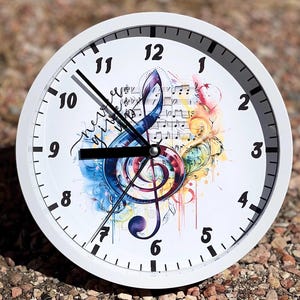 May include: White round clock with black hands and a colorful treble clef design. The clock face has black numbers and a white background. The treble clef is surrounded by musical notes and a watercolor effect.