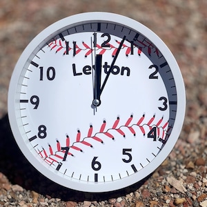 May include: White clock with black numbers and hands. The clock face has a baseball design with red stitching and the name "Leiton" printed on it.