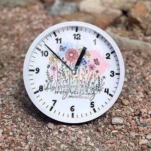 May include: White round clock with black numbers and hands. The clock face features a watercolor background with flowers and butterflies. The text "His mercies are new every morning" and "Lamentation 3:23" are printed on the clock face.