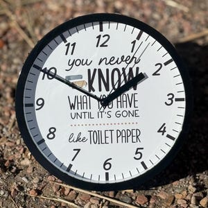 May include: A black and white round clock with black hands. The clock face has the text "You never know what you have until it's gone like toilet paper".