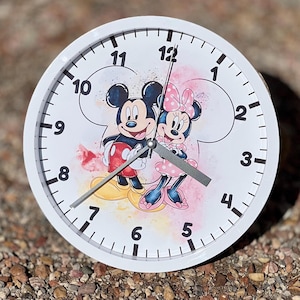 May include: White round clock with Mickey Mouse and Minnie Mouse in the center. The clock face has black numbers and hands. The background is a watercolor design with pink and yellow colors.