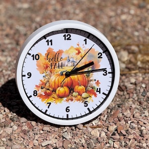 May include: A round white analog wall clock featuring a watercolor illustration of orange pumpkins, autumn leaves, and a small building. The clock face displays the text "hello FALL" in black script and includes black hour, minute, and second hands.