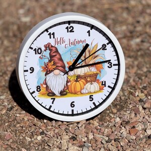 May include: A round white wall clock with a black frame and black hands. The clock face features a gnome with a long white beard and a brown hat, surrounded by pumpkins, corn, and autumn leaves, with the text Hello Autumn printed in script.