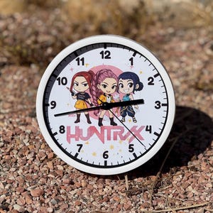 May include: A white-framed wall clock with a pink face featuring three cartoon figures and the text "HUNTRXX." The clock hands point to approximately 8:18. The clock is set on a bed of small, reddish-brown pebbles.