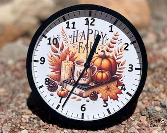 May include: A black and white round clock with a white face. The clock face features a fall-themed design with a book, a mug of coffee, a candle, and pumpkins. The clock face also has the text "Happy Fall" in black lettering.