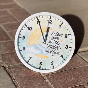 May include: White clock with black hands and numbers. The clock face has a watercolor background with a yellow crescent moon, white clouds, and stars. The text on the clock face reads "I love you to the moon and back".