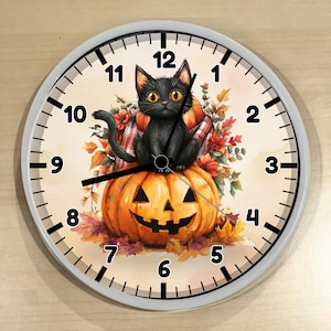 May include: A round wall clock with a white frame and a light-colored face featuring a black cat sitting on a carved orange jack-o'-lantern. The design includes autumn leaves and a plaid scarf, with black numbers from 1 to 12 and black clock hands.