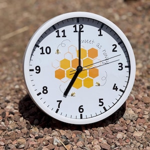 May include: White clock with black hands and a honeycomb design. The clock face says "Sweet as honey".