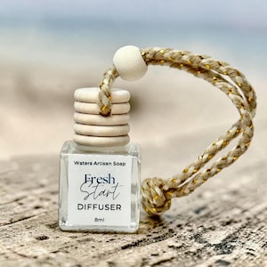 May include: A small clear glass 8ml diffuser bottle with a ribbed wooden cap and a beige braided cord featuring gold metallic threads. The white label displays the text Waters Artisan Soap, Fresh Start, DIFFUSER, and 8ml in black lettering.