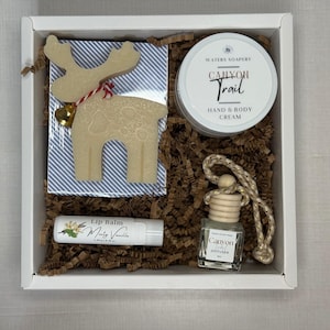May include: A white gift box filled with brown crinkle paper, containing a jar of Canyon Trail hand and body cream, a lip balm, and a small diffuser. Decorative reindeer-shaped soaps and a blue and white striped coaster are also included.