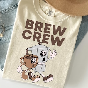 May include: Cream-colored t-shirt with the words "BREW CREW" in brown block letters. Below the text is a cartoon illustration of a coffee cup and a stovetop espresso maker, both with animated faces and limbs.
