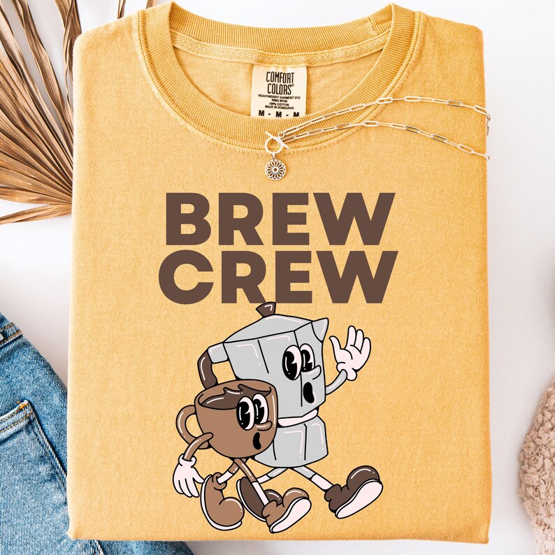 May include: Mustard yellow t-shirt with the words "BREW CREW" in brown block letters. The shirt features a cartoon illustration of a coffee cup and a stovetop espresso maker walking together. A gold necklace is draped across the shirt.