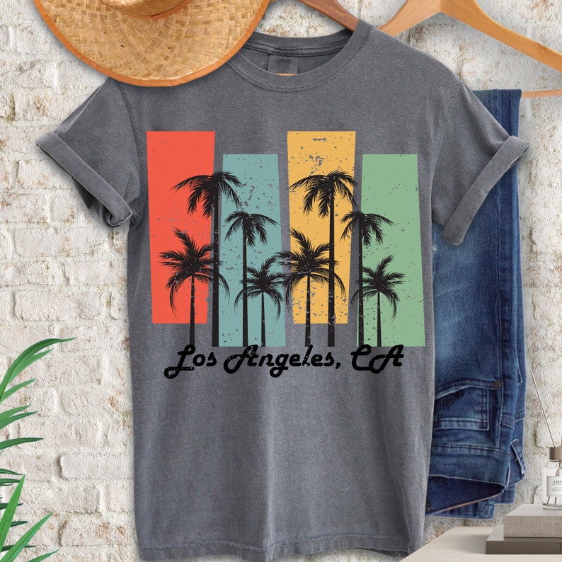 May include: A gray t-shirt with a vintage-style graphic of palm trees in front of colorful stripes. The text "Los Angeles, CA" is printed below the graphic.