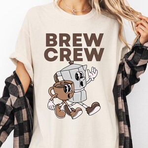 May include: Cream-colored t-shirt with the words "BREW CREW" in brown block letters. Below the text is a cartoon illustration of a coffee cup and a stovetop espresso maker, both with animated faces and limbs.