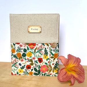 Puede incluir: Un diario cubierto de lino con un forro de tela floral y una etiqueta de madera que dice "Finley". El diario tiene un cierre de bot&oacute;n y un dise&ntilde;o floral con flores rojas, amarillas y rosas.
