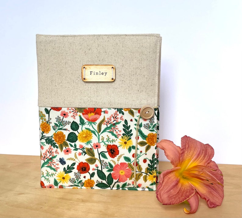 Puede incluir: Un diario cubierto de lino con un forro de tela floral y una etiqueta de madera que dice "Finley". El diario tiene un cierre de bot&oacute;n y un dise&ntilde;o floral con flores rojas, amarillas y rosas.