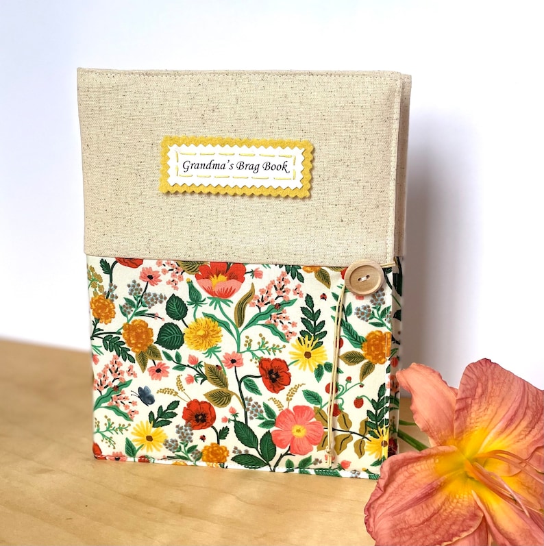 Puede incluir: Una cubierta de libro de lino beige con una etiqueta amarilla que dice "Grandma's Brag Book". La cubierta tiene un patr&oacute;n floral con flores rojas, amarillas y rosas. Un bot&oacute;n de madera est&aacute; unido a la cubierta.