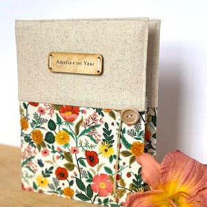 Puede incluir: Un &aacute;lbum de recortes de lino beige con una cubierta de tela floral y una etiqueta de madera que dice "Amelia's 1st Year".