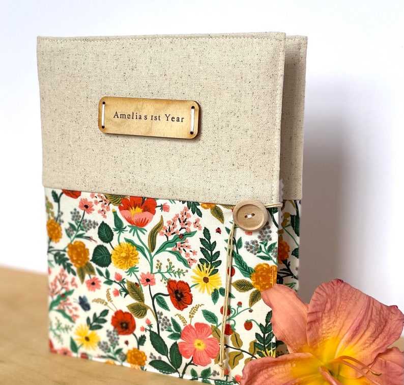 Puede incluir: Un &aacute;lbum de recortes de lino beige con una cubierta de tela floral y una etiqueta de madera que dice "Amelia's 1st Year".