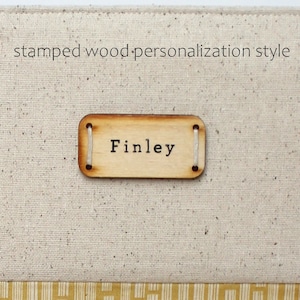 Puede incluir: Primer plano de una etiqueta de madera con el nombre "Finley" estampado en ella. La etiqueta est&aacute; unida a una superficie de tela beige con una cuerda blanca.