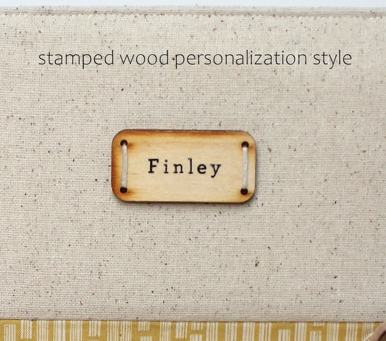Puede incluir: Primer plano de una etiqueta de madera con el nombre "Finley" estampado en ella. La etiqueta est&aacute; unida a una superficie de tela beige con una cuerda blanca.
