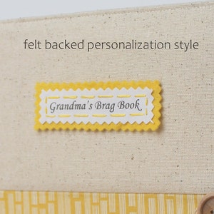 Puede incluir: Una etiqueta de fieltro amarilla con un borde festoneado y costuras blancas. La etiqueta dice "Grandma's Brag Book" en letra cursiva negra.