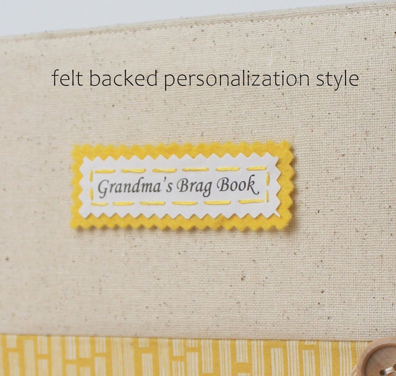 Puede incluir: Una etiqueta de fieltro amarilla con un borde festoneado y costuras blancas. La etiqueta dice "Grandma's Brag Book" en letra cursiva negra.