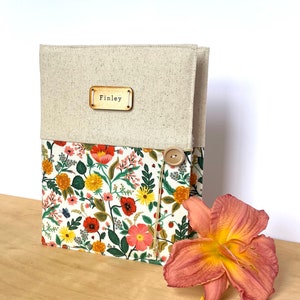 Puede incluir: Una cubierta de diario personalizada de lino y tela floral con una etiqueta de nombre de madera que dice "Finley" y un cierre de bot&oacute;n.