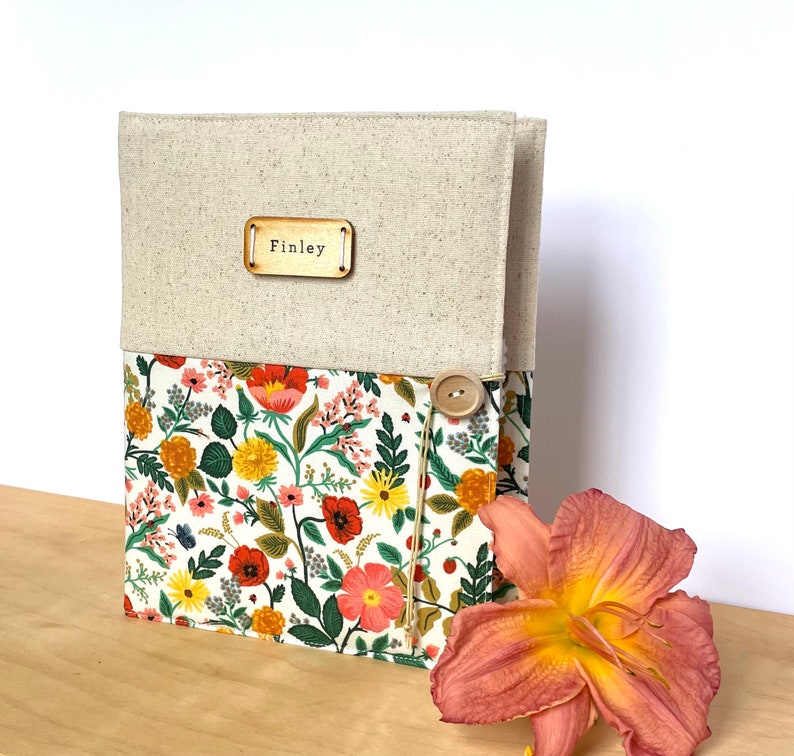 Puede incluir: Una cubierta de diario personalizada de lino y tela floral con una etiqueta de nombre de madera que dice "Finley" y un cierre de bot&oacute;n.