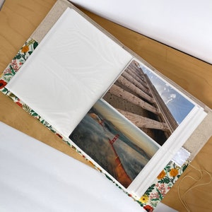 Puede incluir: Un &aacute;lbum de fotos de tela blanca con un patr&oacute;n floral en la portada. El &aacute;lbum est&aacute; abierto para revelar dos postales. Una postal muestra el Golden Gate Bridge en San Francisco, y la otra muestra una fila de columnas.