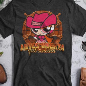 Puede incluir: Camiseta gris oscuro con un personaje de dibujos animados con sombrero y atuendo de vaquero rosa, sobre un diseño de rueda de madera. El texto "ANTES MUERTA que sencilla" se muestra en letras doradas.