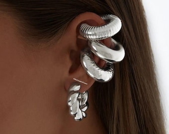 May include: Silver ear cuff earrings with a layered design. The ear cuff is made of three different sized hoops, each with a smooth, polished finish. The earrings are worn on the upper part of the ear.