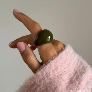 Könnte beinhalten: Olivgrüner Harzring mit glattem, abgerundetem Design. Der Ring ist einfarbig und hat eine glänzende Oberfläche. Der Ring wird am Finger getragen.