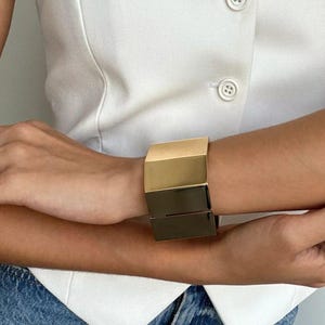 Könnte beinhalten: Ein geometrisches Armreif-Armband in Gold und Schwarz. Das Armband besteht aus zwei Teilen, einem goldenen und einem schwarzen, die miteinander verbunden sind.