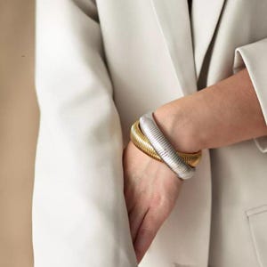 以下が含まれることがあります： リブ模様のゴールドとシルバーのブレスレットが2つ重ねられています。ブレスレットは手首につけられています。