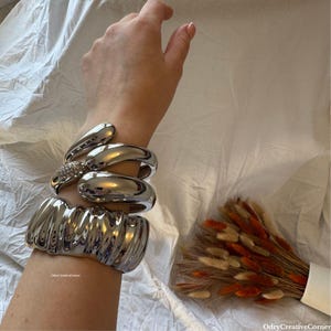 Puede incluir: Un brazalete de plata con un diseño texturizado. El brazalete está hecho de múltiples bandas que se apilan una encima de la otra.