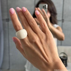 Op de afbeelding: Een grote, crèmekleurige, dikke ring wordt aan een vinger gedragen. De ring is glad en afgerond. De hand heeft roze nagellak. De achtergrond is een wazige spiegelreflectie.