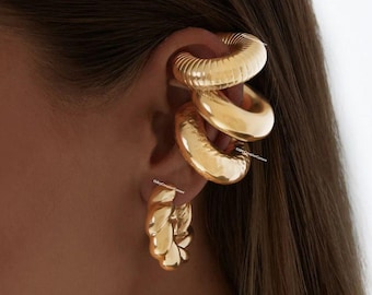 May include: Gold ear cuff earrings with a twisted design. The ear cuff is made of three stacked rings, each with a different texture. The earrings are worn on the upper part of the ear.
