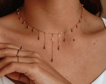May include: A gold chain necklace with small round beads hanging from the chain. The necklace is a choker style and is worn by a person.