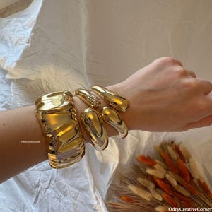 Pode incluir: Um conjunto de três pulseiras de metal dourado. As pulseiras estão empilhadas no pulso de uma pessoa. As pulseiras são de diferentes formas e tamanhos. A pulseira de baixo é texturizada e as duas pulseiras de cima são lisas.
