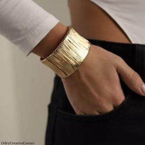 May include: A gold-toned bracelet with a textured, vertical stripe design. The bracelet is worn on a person's wrist.