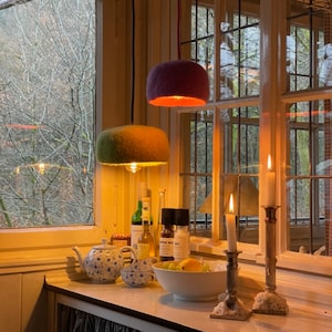 Könnte beinhalten: Zwei Hängelampen mit flauschigen violetten und grünen Schirmen beleuchten eine Küchentheke mit Kerzenständern, einer Schüssel mit Obst und einem Teekannen- und Milchkännchen-Set.