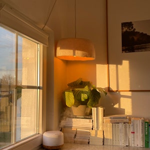 Könnte beinhalten: Eine weiße, flauschige, runde Leuchte hängt von der Decke in einem Raum mit einem Fenster, einer Pflanze und einem Bücherregal voller Bücher. Die Leuchte strahlt ein warmes Licht in den Raum.