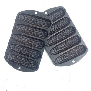 May include: Two black cast iron cornbread molds with six individual cornbread molds on each. The molds are on a white surface.