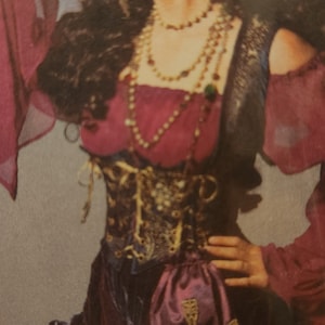 May include: A woman wearing a red and black costume with a gold and black corset, a purple sash, and a gold beaded necklace. She is wearing a red headscarf and has long dark hair.