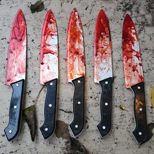 May include: Five kitchen knives arranged in a row, each featuring a silver blade covered in red paint splatters resembling blood. The knives have black handles secured with three silver rivets, presenting a uniform design across all five items.